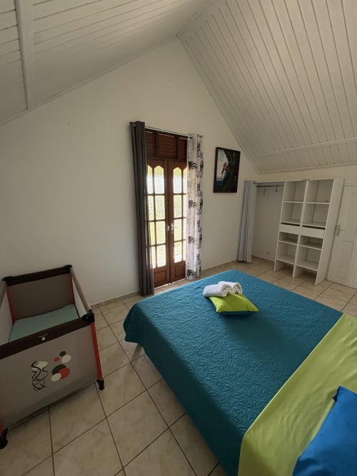 Chambre lumineuse de la villa L'ASETA PARADISE à Grand-Bourg avec lit bleu, parc pour bébé, portes en bois, étagères blanches et carrelage