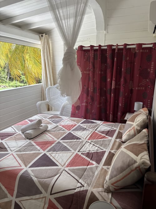 Chambre spacieuse de la villa L'Aseta Paradise avec parure patterned, rideaux bordeaux, fauteuil blanc et vue sur le jardin