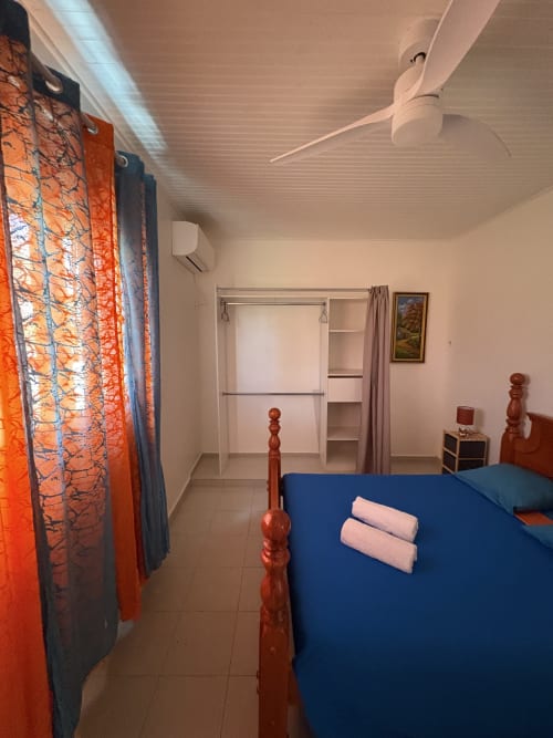Chambre spacieuse de la villa L'ASETA PARADISE avec lit bleu, poteaux en bois, ventilateur blanc et rideaux orange