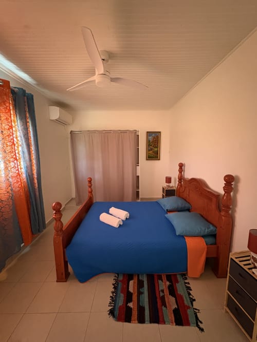 Chambre spacieuse de la villa L'Aseta Paradise à Grand Bourg avec lit bleu, tête de lit en bois, ventilateur de plafond et vue sur le jardin