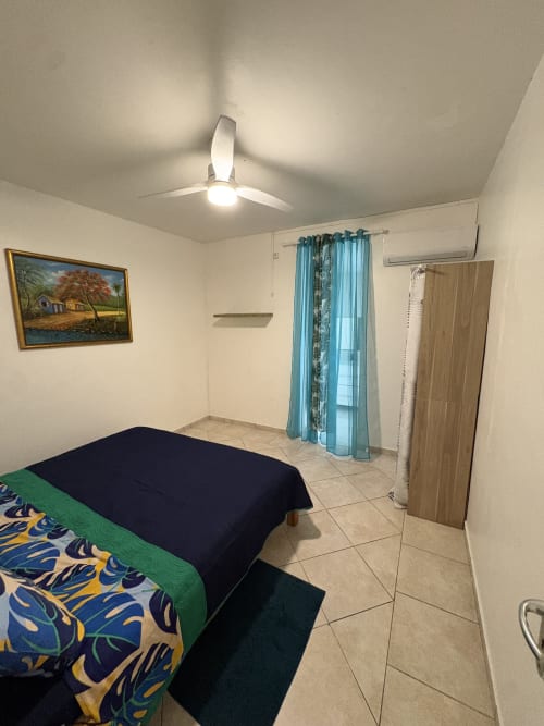 Chambre lumineuse de la villa L'Aseta Paradise, Grand Bourg Guadeloupe avec ventilateur de plafond, lit, rideaux turquoise et vue sur le jardin