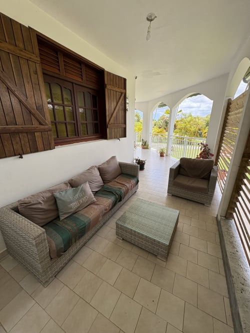 Véranda spacieuse et couverte de la villa L'ASETA PARADISE en Guadeloupe avec mobilier en rotin et vue sur le jardin