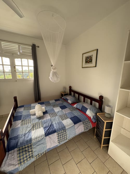 Chambre lumineuse avec parure de lit écossaise bleue, lit en bois, moustiquaire en baldaquin et vue sur le jardin à L'ASETA PARADISE