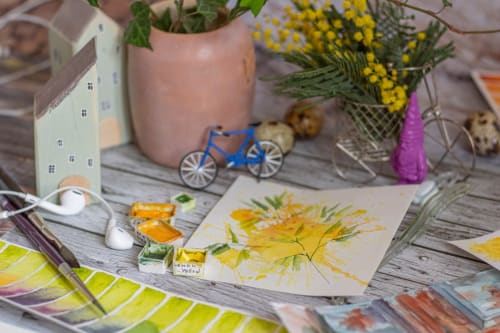 Espacio creativo del Yasi Hostel Lyon con flores pintadas, bicicleta de juguete azul, plantas en macetas y suministros artísticos sobre madera