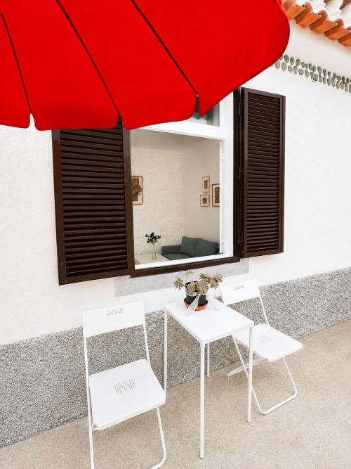 Guarda-sol vermelho oferecendo sombra para cadeiras brancas e mesa na Villas Simone Porto Santo, com venezianas e área de descanso litorânea