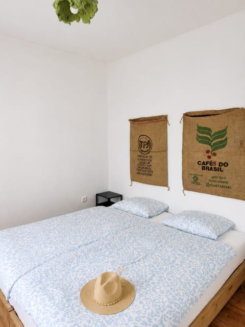 Quarto luminoso nas Villas Simone com roupa de cama azul estampada, arte de parede em sacos de café de juta, chapéu de palha e decoração de grinalda verde