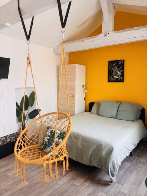 Chambre chaleureuse au Chez Mary Studios avec fauteuil macramé jaune suspendu, lit aux coussins verts, mur d'accent orange et plante en pot