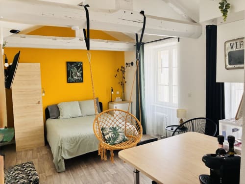 Petit gîte aux thèmes tropicaux à Gannat, mur jaune, chaise suspendue, lit queen-size et espace de vie ouvert