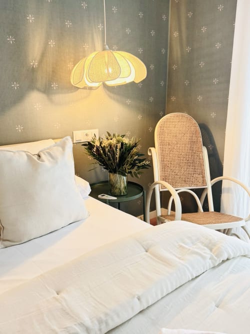 Dormitorio luminoso con lámpara colgante amarilla, ropa de cama blanca, silla de ratán, flores secas y papel tapiz con patrón de copos de nieve