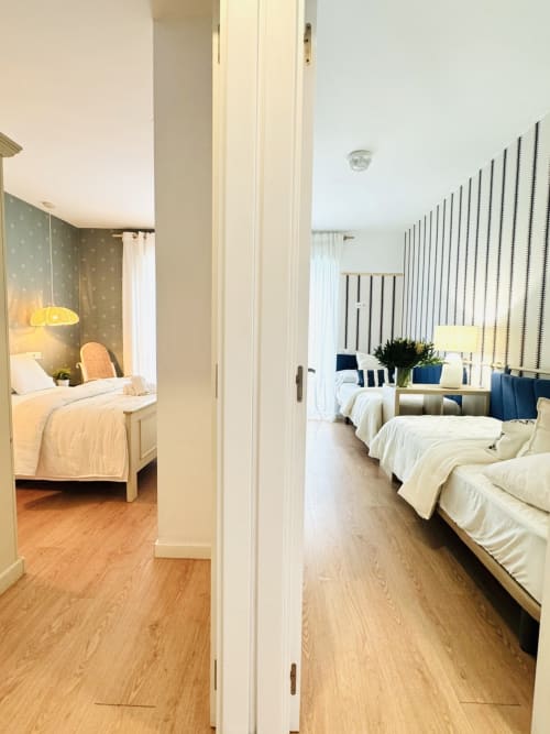 Puerta abierta conectando dos luminosos dormitorios en dúplex de Sinese Design Homes Málaga con camas individuales, papel pintado a rayas
