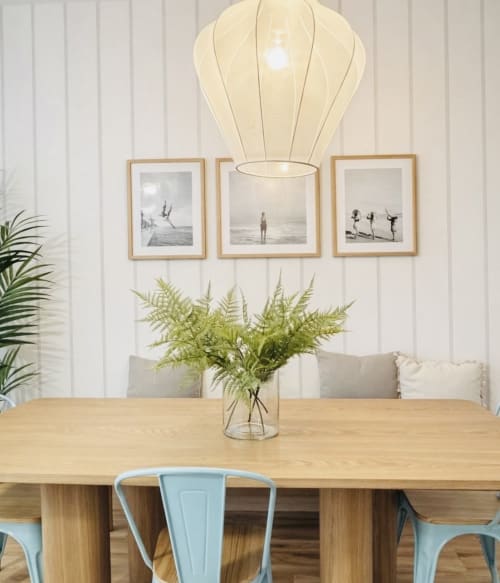 Comedor moderno con mesa de madera, sillas azul claro, lámpara colgante color crema y obra costera enmarcada en la playa de Málaga