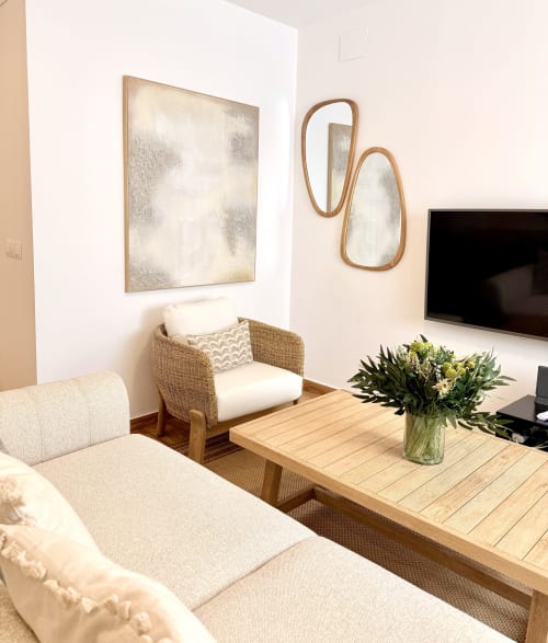 Sala de estar moderna en dúplex frente al mar en Málaga con sofá crema, silla de ratán, mesa de madera y espejos contemporáneos