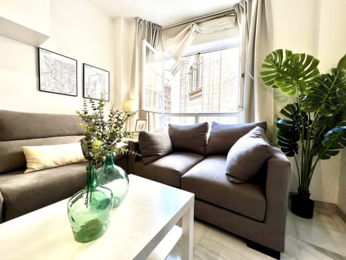 Moderna sala de estar del apartamento Deluxe Catedral de Málaga con sofás grises, plantas y vistas del hito histórico