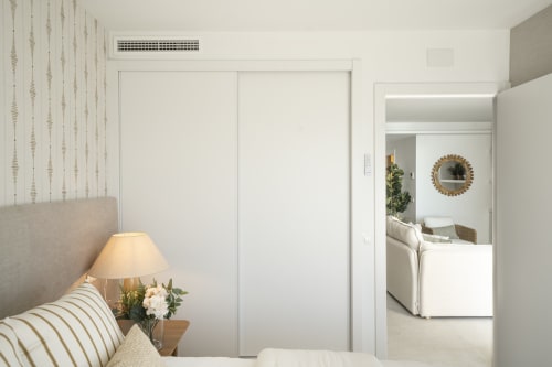 Moderno dormitorio en apartamento Premium ROCA Beach Torremolinos con decoración blanca, lámpara de noche y vista hacia la sala de estar
