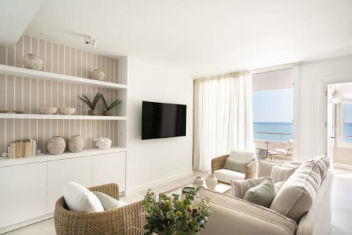 Luminoso apartamento moderno con vistas al mar, estanterías blancas, sofá beige, sillas de mimbre y acceso a la playa