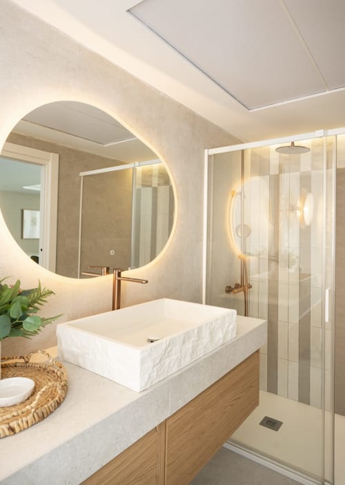 Baño moderno con espejo circular retroiluminado, lavabo de piedra blanca y ducha con cristalería en ROCA Beach Torremolinos Premium