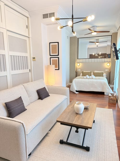 Moderno estudio en Málaga con sofá beige, mesa de madera y dormitorio visible tras mampara de cristal