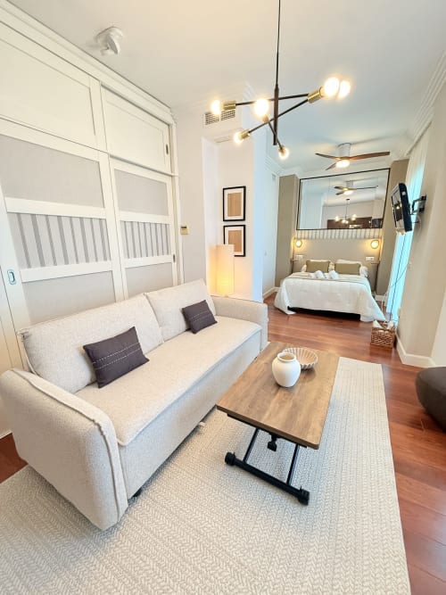 Moderno apartamento estudio en Málaga con sofá beis, mesa de café de madera y dormitorio visible a través de puerta abierta