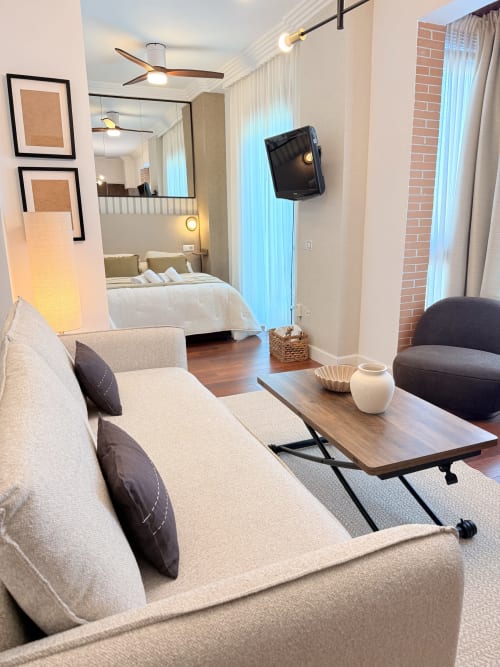 Moderno estudio en Málaga con sofá beige, mesa de madera, TV de pantalla plana y dormitorio con ropa de cama blanca visible