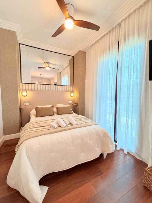 Dormitorio moderno en apartamento de Málaga con cama queen, ventilador de techo, pared con cabecero espejado, suelos de madera y luz natural