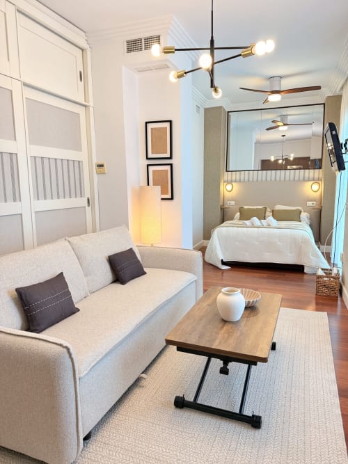 Moderno estudio en el centro de Málaga con sofá beige, mesa de café de madera y dormitorio con cabecero espejado