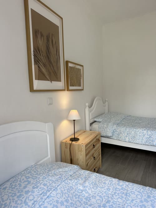 Quarto duplo em Villas Simone com colchas azuis estampadas, criado de madeira, abajur e arte botânica na parede.