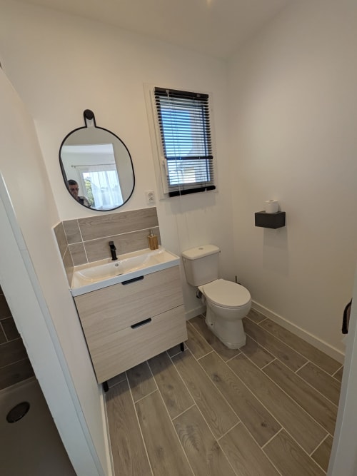 Salle de bains moderne à la Résidence des dunes avec lavabo, miroir, toilettes, carrelage imitation bois et store à fenêtre