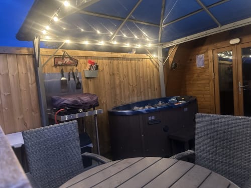 Dragonfly Pod hot tub terrace at dusk with glowing pergola lights, seating area, and wooden cabin at Acorns Glamping,
