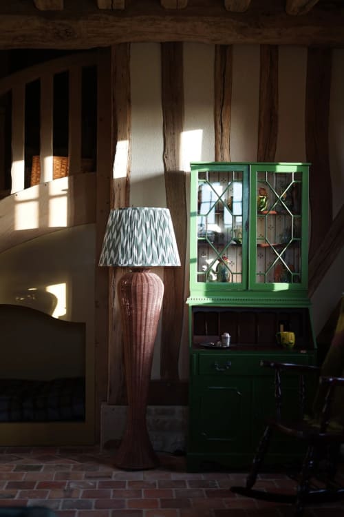 Intérieur rustique de la Ferme La Roquerie avec poutres apparentes, lampe rose, armoire verte et lumière naturelle des fenêtres
