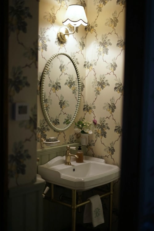Salle de bains élégante à la Ferme La Roquerie avec papier peint fleuri, miroir rond, lavabo sur colonne et applique murale