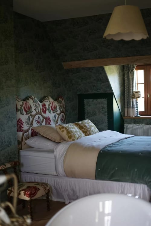 Chambre douillette de la Ferme La Roquerie avec tête de lit fleurie, coussins décoratifs, suspension et vue sur la campagne