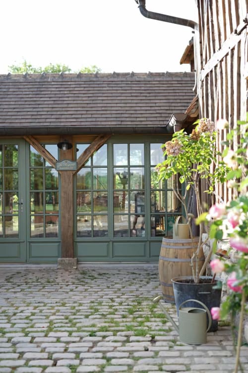 Cour du Mas de la Roquerie avec portes vertes, poutres en bois, fleurs en pots et vues champêtres