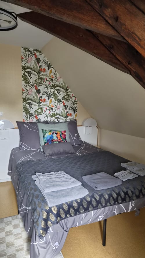Chambre douillette sous les combles à l'Hôtel Le Perroquet Bleu avec papier peint tropical, literie sombre, poutres apparentes et serviettes pliées