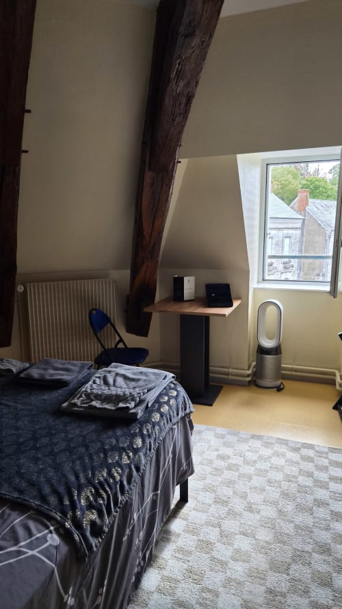 Cozy attic bedroom at Hôtel Le Perroquet Bleu with dark wooden beam, desk with laptop, air purifier, and valley view window.