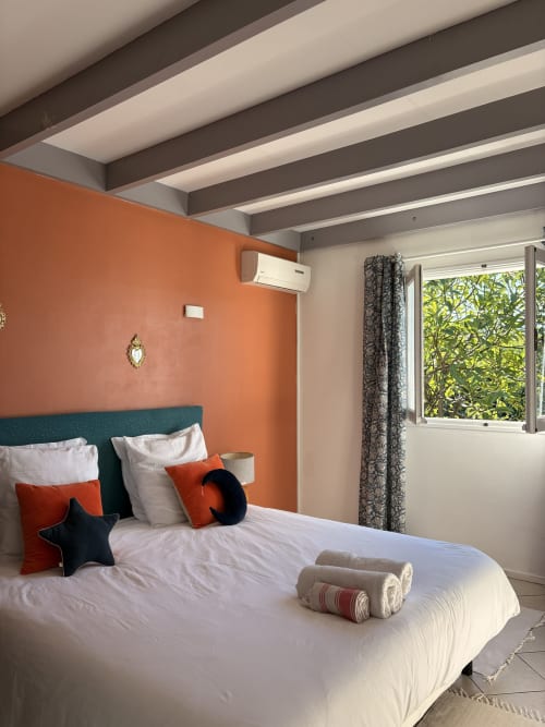 Modern bedroom at La Villa Maraloupia with orange accent wall, teal headboard, white bedding, and ocean views through window