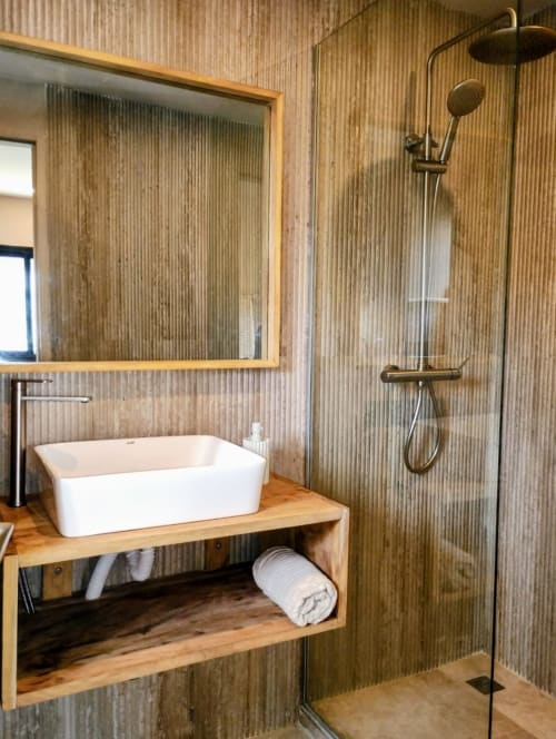 Salle de bain moderne à Nomads Togo avec lavabo blanc, meuble en bois, douche à effet pluie et murs en tôle ondulée à Lomé
