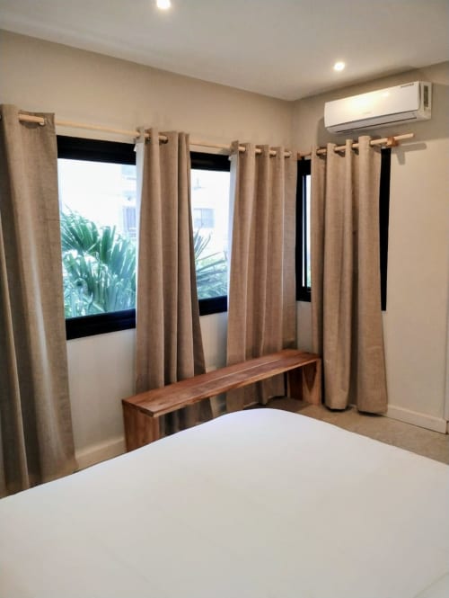 Chambre aux Nomads Togo avec rideaux beige, banc en bois, climatiseur et vue sur le jardin à Lomé