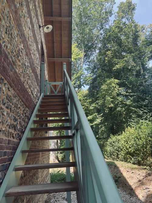 Escalier en bois avec rampes vertes menant à l'entrée de la maison de pierre du Manoir de Daubeuf, entouré de verdure