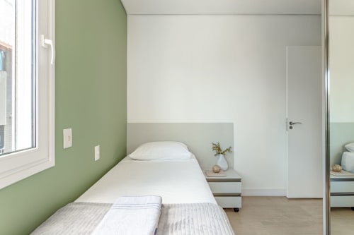 Quarto moderno com parede de destaque verde, cama branca, criado com decoração e espelho no apartamento de Canasvieiras, Florianópolis