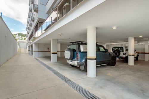 Garagem de estacionamento coberta no Sua Estadia SC em Florianópolis com SUV azul, pilares de suporte e edifício residencial acima