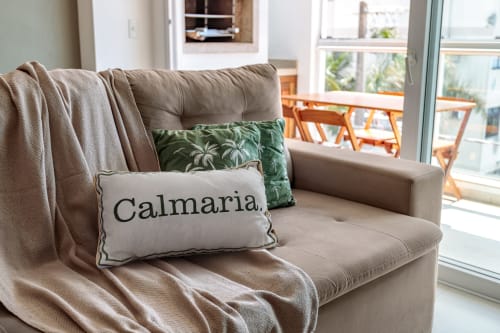 Sofá bege com almofadas decorativas estampadas com 'Calmaria' e padrão de palmeiras verdes, manta sobre o braço, janelas com vista para o mar