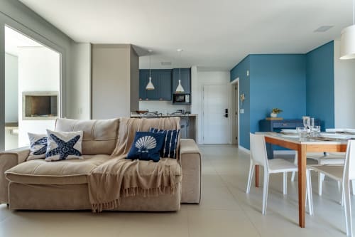 Apartamento moderno à beira-mar em Florianópolis com sala aberta, sofá bege, parede azul-teal, mesa de madeira, e