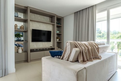 Sala moderna em tons bege com painel de TV, sofá creme, manta estampada e vista para o oceano em Canasvieiras, Florianópolis