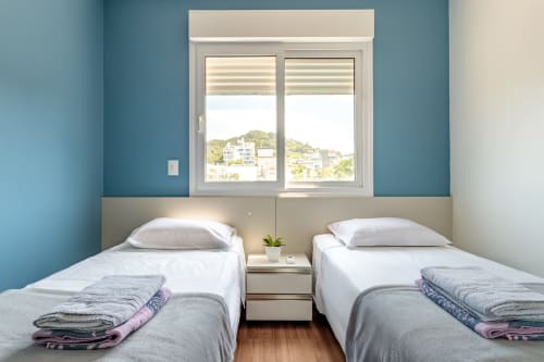 Camas gêmeas em quarto moderno com vista para o mar, apartamento na praia de Florianópolis, paredes azul-petróleo, janela com persianas