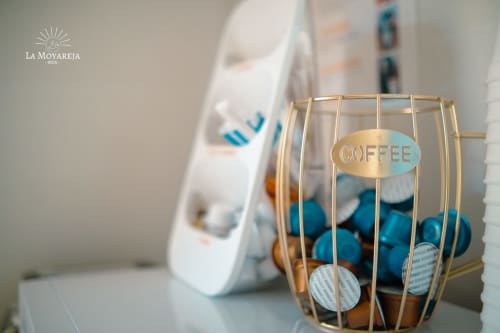 Cesta de alambre dorado con cápsulas de café azules etiquetadas 'COFFEE' en Hostal La Moyareja en Rota