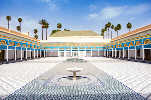 Cour du Riad Casa Angelo à Marrakech avec arcades carrelées de bleu, fontaine centrale et palmiers sous un ciel dégagé.