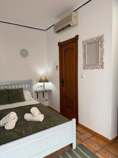 Habitación en el Hotelito Oliva Pensión en Oliva con cama blanca, ropa de cama oscura, lámpara de noche, puerta de madera y espejo mural