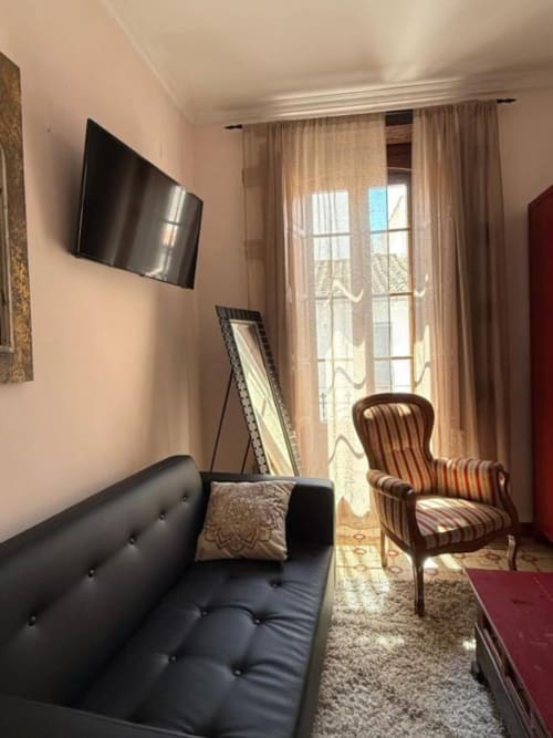 Luminosa habitación del Hotelito Oliva con sofá negro, sillón a rayas, TV de pared y balcón con vista a Oliva