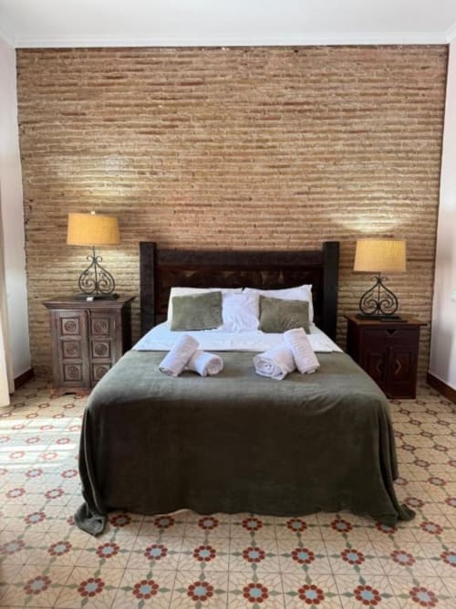 Dormitorio del Hotelito Oliva con cama de madera oscura, pared de ladrillo tostado, lámparas decorativas y suelo de azulejos con patrón cerca de Oliva, España