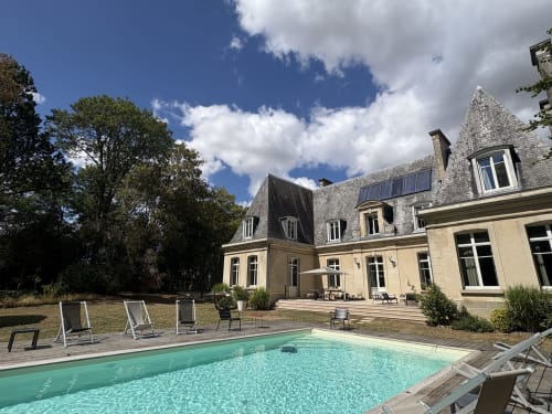 Le château d'Hermonville, gîte de luxe avec piscine turquoise, transats et manoir historique en pierre sous ciel bleu